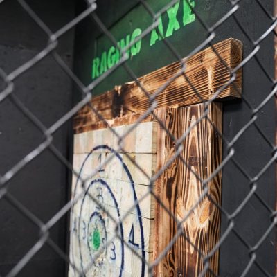 View of an axe throwing target behind a chain-link fence with 'Raging Axe' sign above.