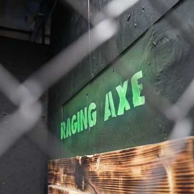 Close-up of stenciled green 'Raging Axe' text on a dark wall with chain-link fence.