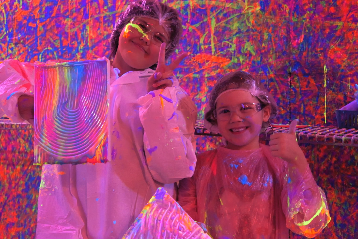 Two kids in protective gear holding neon paint art pieces with splatter background.