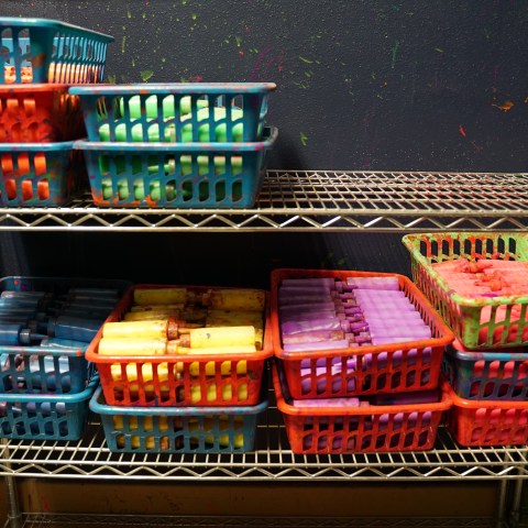 Colorful baskets on metal shelves filled with paint bottles and utensils against a painted wall.