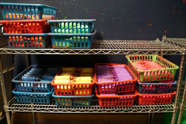 Colorful baskets on metal shelves filled with paint bottles and utensils against a painted wall.