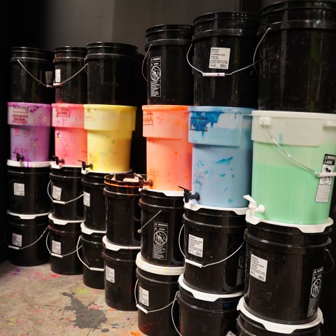Stacked black and colorful paint buckets against a dark wall in a studio.