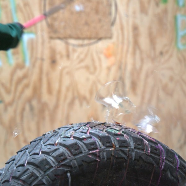 Person in green jacket swings bat at tire with glass shards flying.