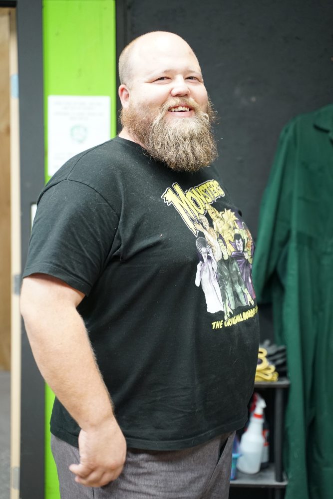 Smiling man with a beard wearing a monsters-themed t-shirt stands indoors with green background.